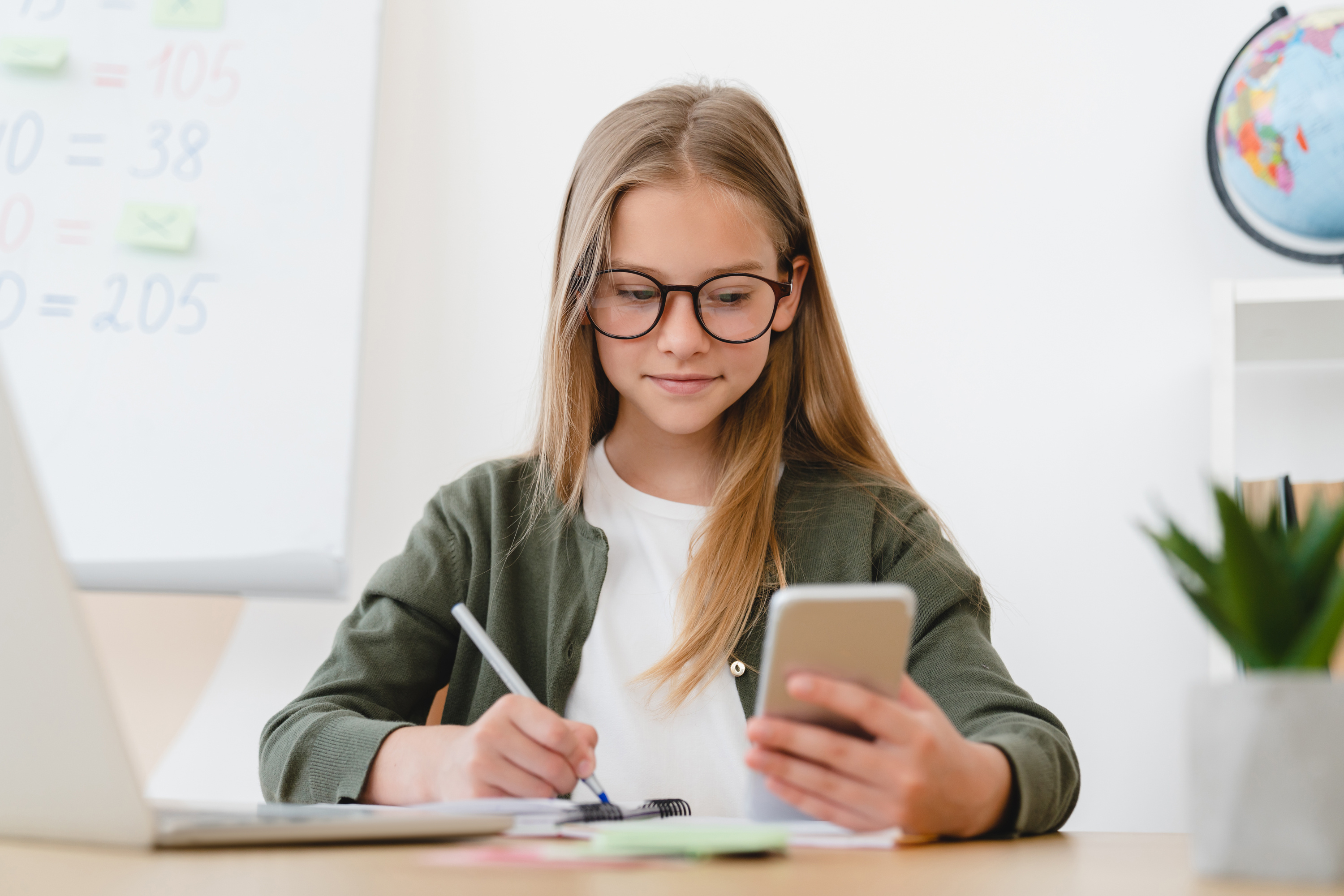 Young girl with glasses, writing notes while looking at a smartphone, with a laptop and a globe in the background.






